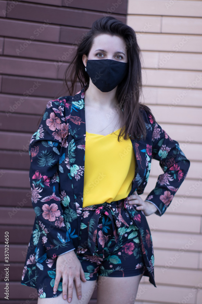 Attractive young brunette woman in yellow t-shirt and flowery shorts costume. In medical black face mask. brown wall background. modern reality. covid-19 concept. copy space. High quality photo