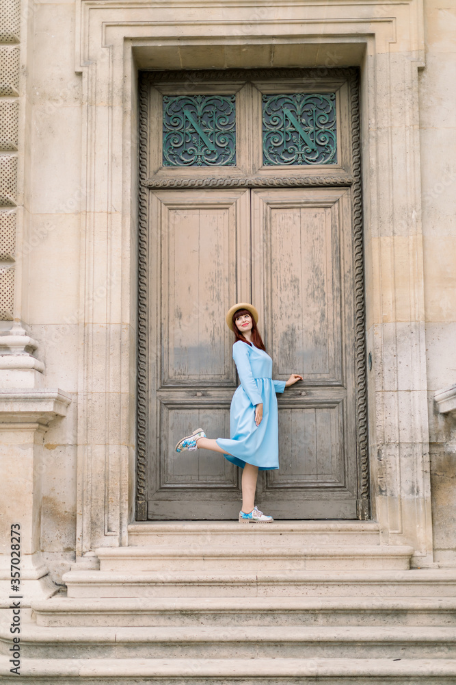 Charming elegant young woman wearing blue dress, posing in front of ancient wooden door of old vintage building with one leg up