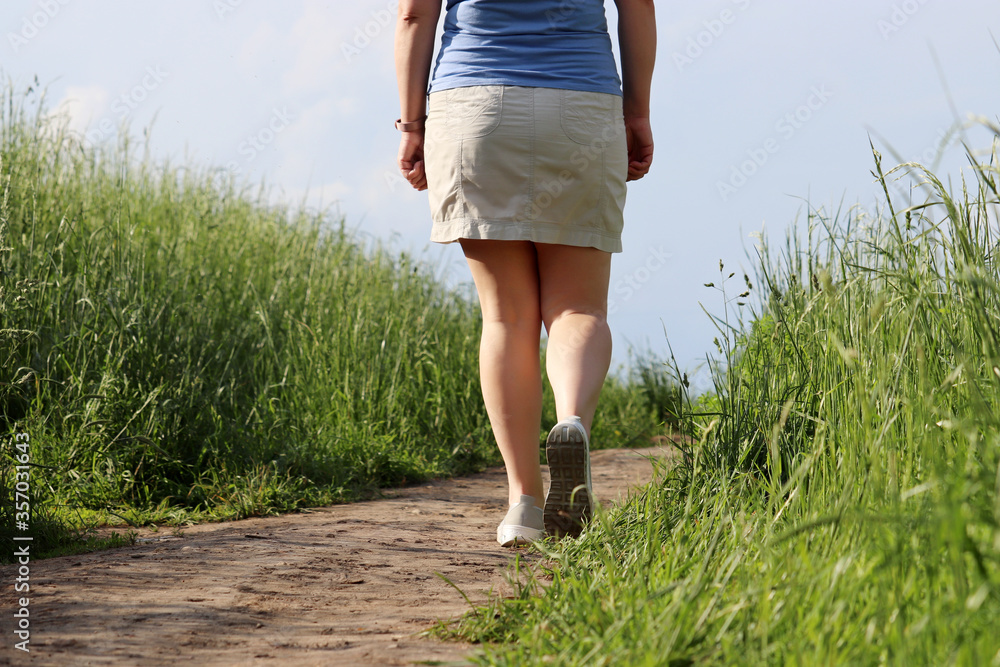 Woman in summer skirt climbs to the top on a path, rear view. Rural scene with meadow   overgrown of high grass on background of blue sky, vacation in countryside