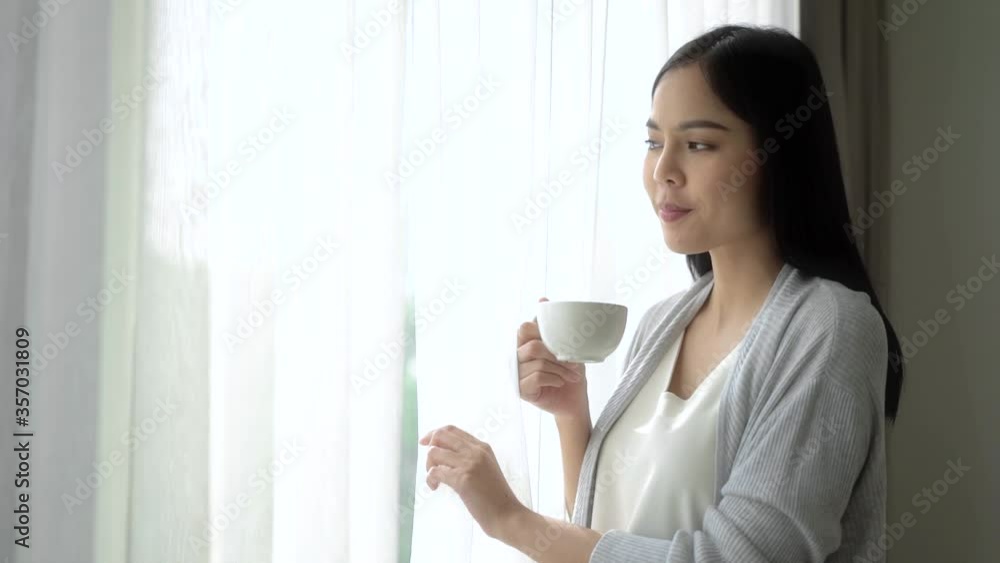 Young asian woman opening curtains looking out window enjoying fresh new day feeling rested and drinking coffee at home. Slow Motion Full Hd. Young asian woman opening curtains looking out window enjoying fresh new day feeling rested and drinking coffee at home. Slow Motion Full Hd.