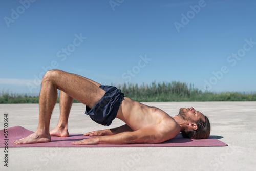 Concept: sport. Attractive blond man with long hair collected with a beard. Doing pilates and yoga outdoors. Pelvic tilt to pelvic curl pose.
