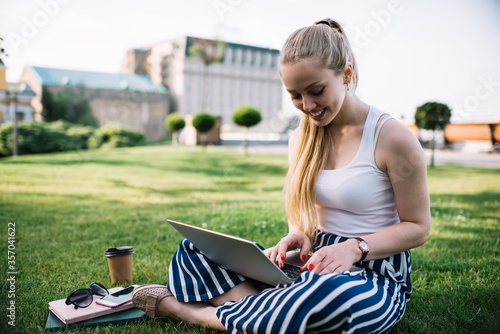 Cheerful Caucasian female freelancer sitting on summer grass 