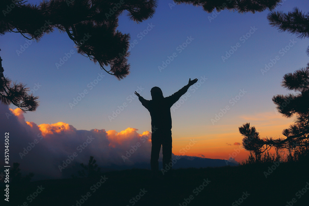 Silhouette of a man in the mountains at sunset in the pines, arms spread, greeting later in the evening Silhouette of a man in the mountains at sunset in the pines, arms spread, greeting later in the evening