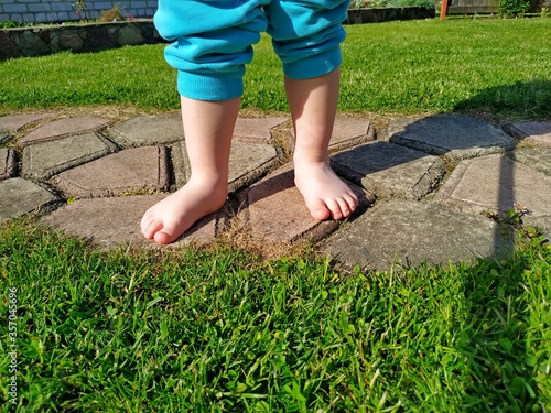 a small child in aqua blue pants and barefoot stands on tile on a green lawn.