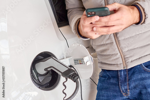 Man charging electric automobile and using his smartphone for payment at charging point.