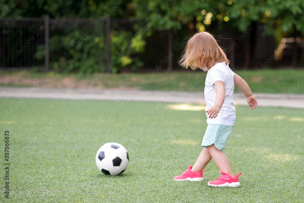 Toddler girl in pink sneakers playing football at stadium. Little blonde child ready to kick soccer ball at green field outdoors. Summer activities for children and future athlete concept. Copy space