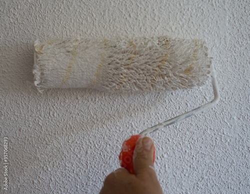 Close-up of paint roller painting a white wall. The wall has popcorn ceiling texture. Home renovation project photo