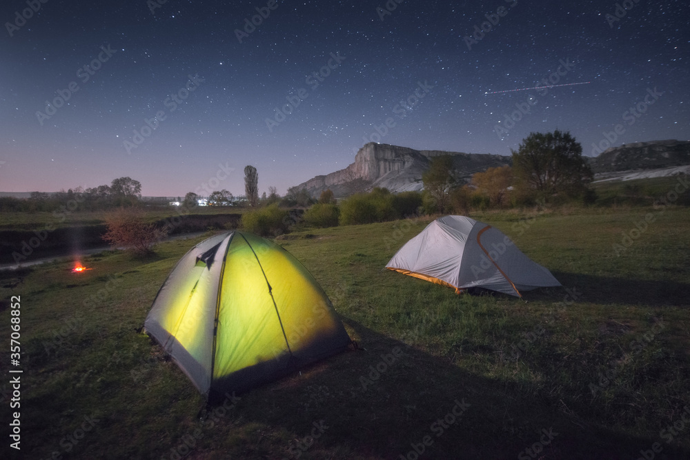 Picturesque night landscape with two tourist tents. The green valley of a mountain river with a beautiful White Rock. Campground in the meadow. Summer starry sky, Perseid meteor.