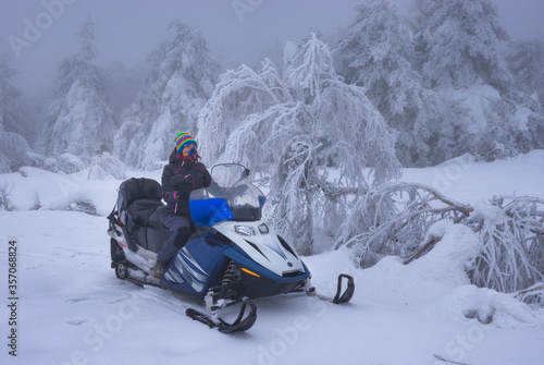 oung girl in a multi-colored hat rides a snowmobile through a fabulous misty winter forest covered with fluffy freshly fallen snow. Tourism, travel, winter activities concept. 
