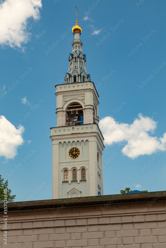 The high white bell tower of the Orthodox Church with a golden cross and a tower clock surrounded by a high stone fence