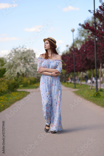 A beautiful girl in a blue dress and a straw hat walks along an empty Boulevard in the city garden. in summer
