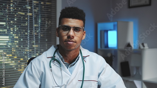 Close-up of young african doctor looking to the camera and telling about something in the evening. He working home with skyscrapers in the background.