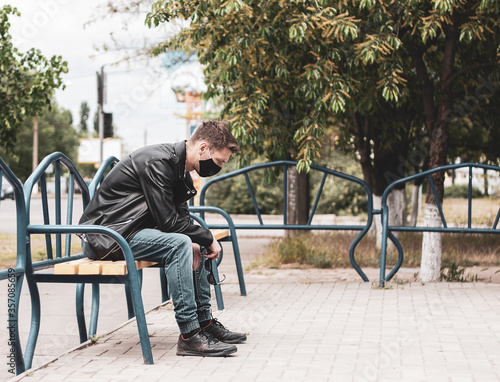Caucasian men sits on the bench and looks straight away. He is depressed. Dressed into jeans, T-shirt, leather jacket and black protective mask. (Picture has a preset dark colors.)
