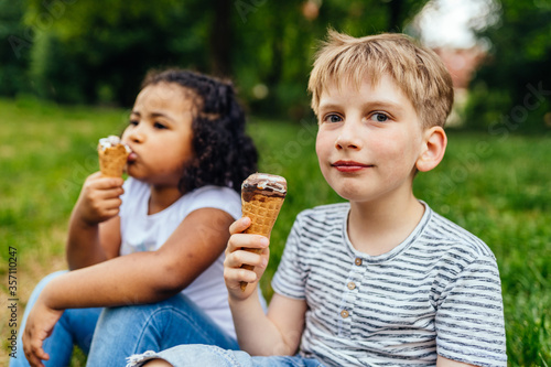 Cute little blond boy and mixed race hispanic little girl eating ice cream in green grass park  close up. Different age multiethnic children enjoying time together.