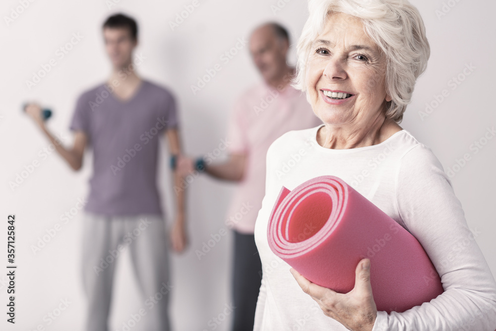 Happy senior woman holding pink joga mat, men exercising in background