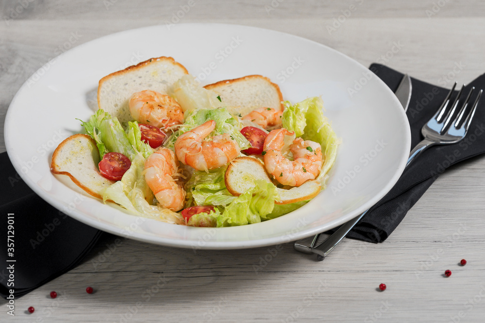 White plate of Caesar salad with shrimps (Ingredients: iceberg Lettuce, crispy croutons, cherry tomatoes, Parmesan cheese and shrimps fried in olive oil) on a light gray background with dark napkins. White plate of Caesar salad with shrimps (Ingredients: iceberg Lettuce, crispy croutons, cherry tomatoes, Parmesan cheese and shrimps fried in olive oil) on a light gray background with dark napkins.