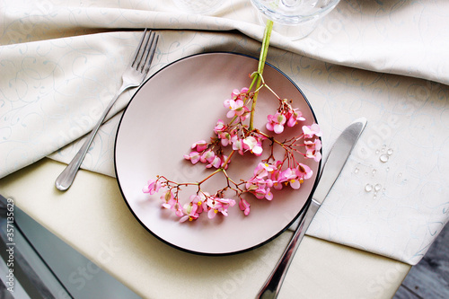 dining table with a beige tablecloth and a plate. table setting with begonia flowers. photo of a table with utensils top view.