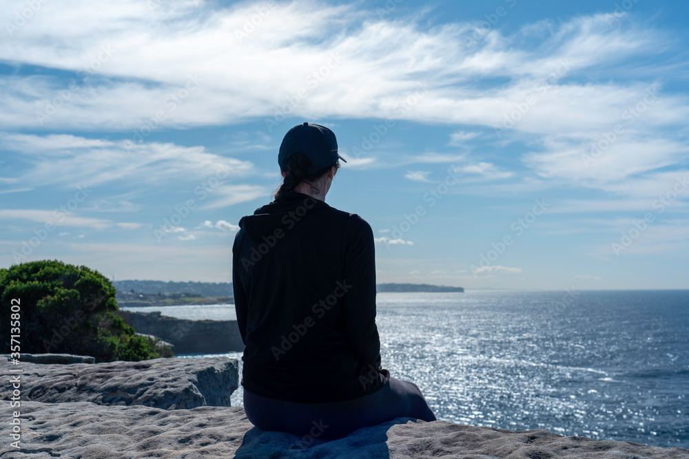 View of lady wearing a black workout t-shirt from behind at the beach overlooking the peaceful blue ocean View of lady wearing a black workout t-shirt from behind at the beach overlooking the peaceful blue ocean