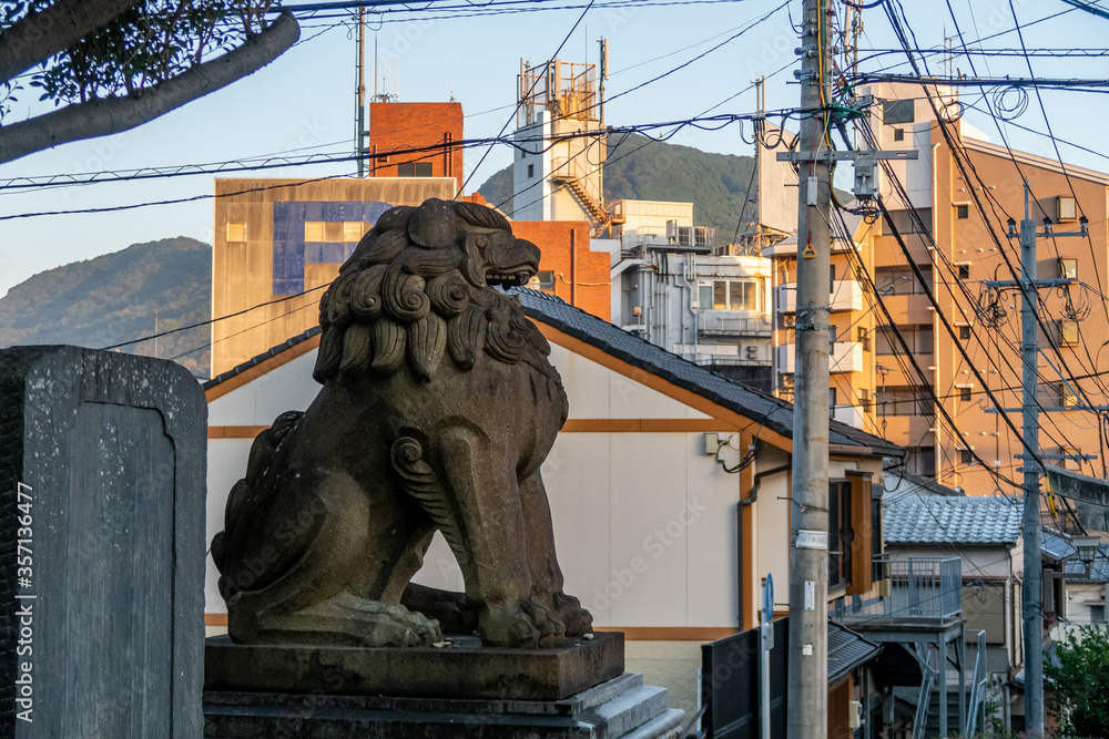 Fototapeta premium Traditional stone lion statue guarding Nagasaki city, Japan, with buildings and electric power cables and poles in the background.