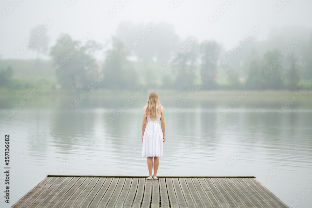 One young woman in white dress standing on edge of footbridge and staring at lake in summer. Mist over water. Foggy air. Thinking about life. Spending time alone in nature. Back view.