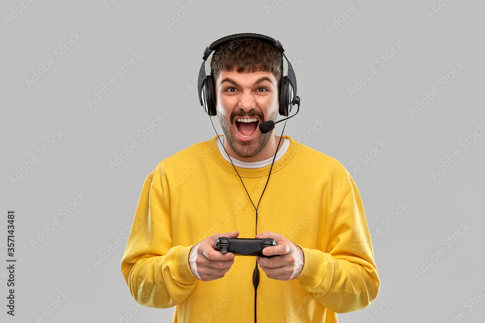 technology, gaming and people concept - angry young man or gamer in headphones with gamepad playing and streaming video game over grey background - obrazy, fototapety, plakaty 