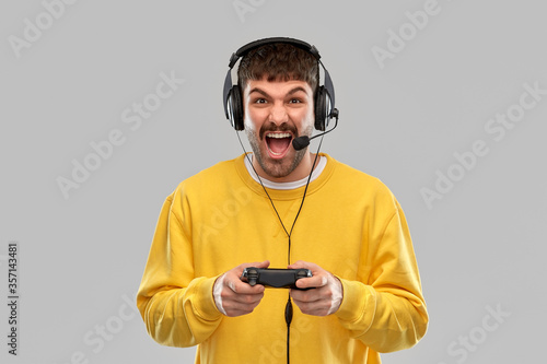technology, gaming and people concept - angry young man or gamer in headphones with gamepad playing and streaming video game over grey background