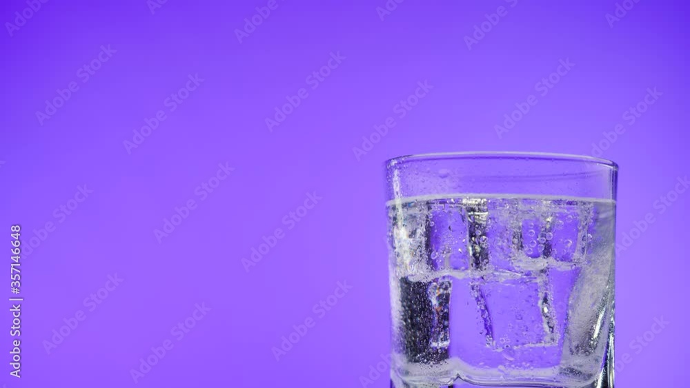 Glass full of cold sparkling water with ice cubes rotate on a violet background. Transparent fizzy drink. Moving bubbles in soda. Refreshing mineral water. Glass full of cold sparkling water with ice cubes rotate on a violet background. Transparent fizzy drink. Moving bubbles in soda. Refreshing mineral water.