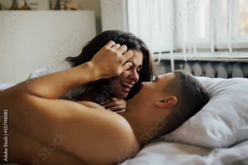Joyful couple in good mood lying in bed at bedroom. Close up photo of a caucasian woman with brown hair lying on her boyfriend. Smiling playful young lady in male white shirt sitting on her happy man 
