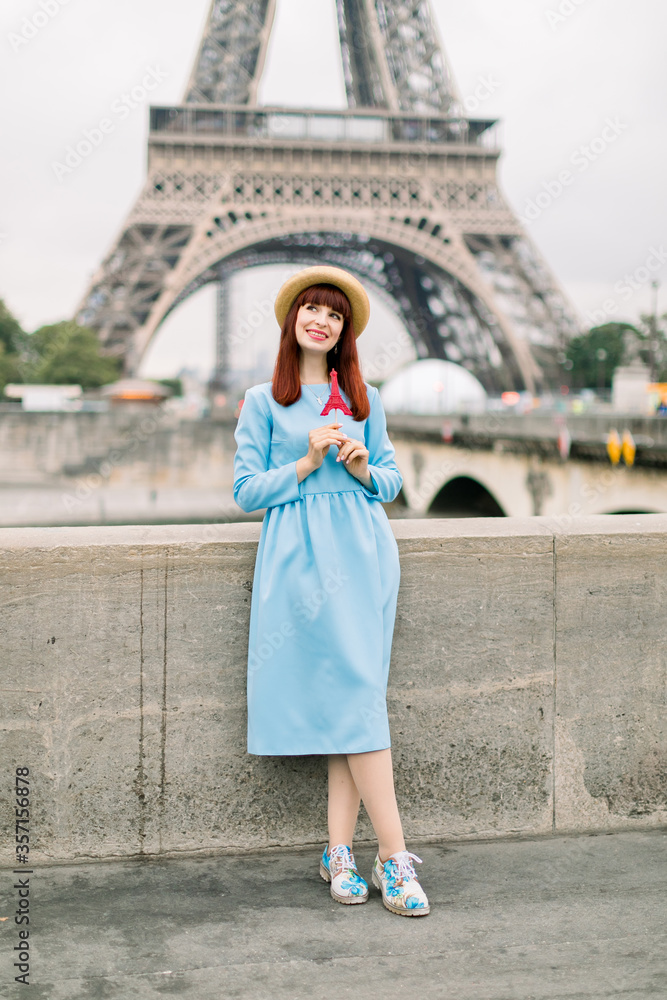 pretty young woman in blue dress and straw hat with red eiffel tour shaped lollipop in hands standing on the background of Eiffel tower in Paris