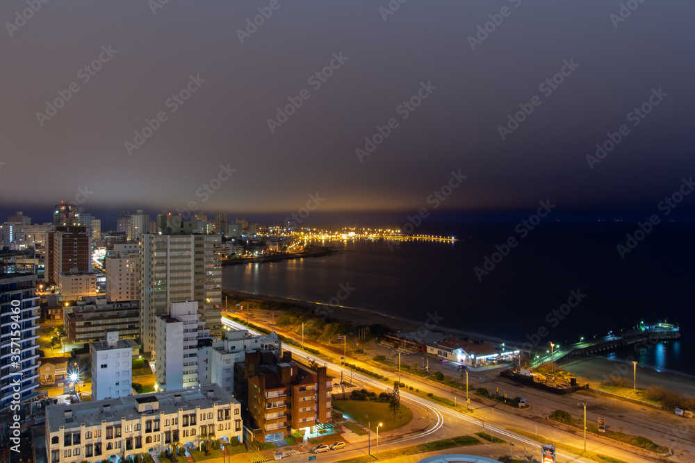 Long exposure panoramic night view of the coastline at a holiday resort in Punta Del Este, Uruguay: the quiet empty beach, the calm water, the car lights passing and the illuminated harbor at the back