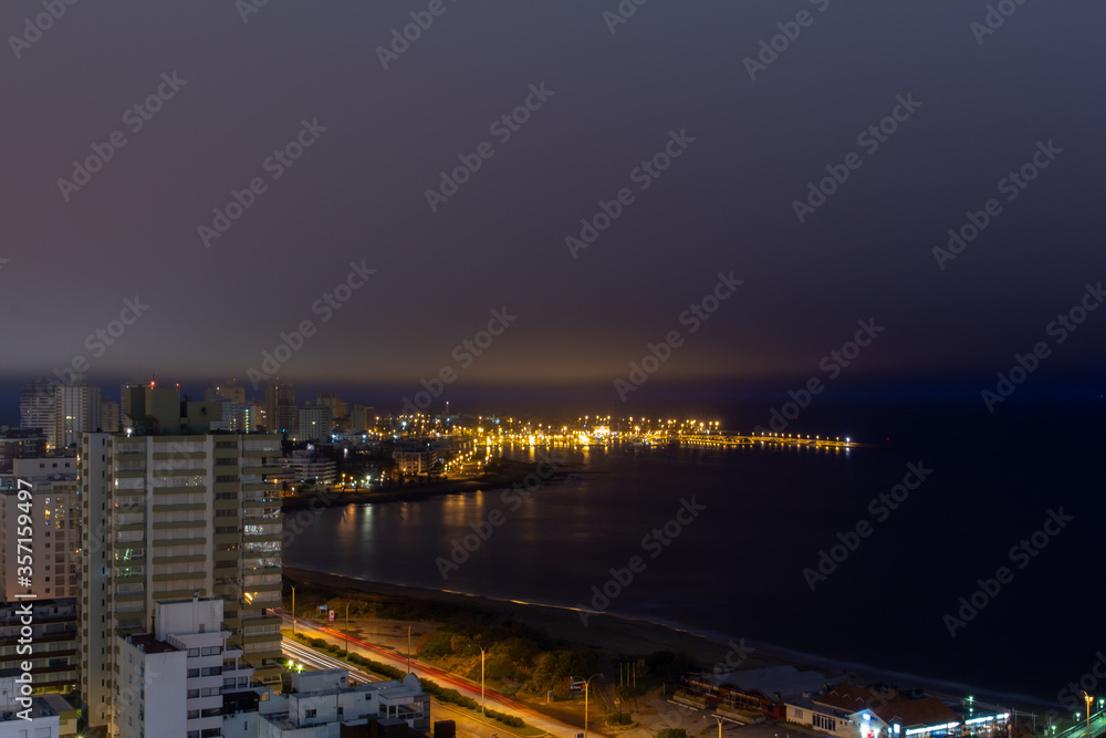 Long exposure panoramic coast view of the illuminated harbor, the calm dark water, the car lights passing and a thick layer of clouds almost covering the buildings at night in Punta Del Este, Uruguay.