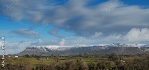 Panorama image of Benbulben flat top mountain in County Sligo, Ireland; Warm day, cloudy sky, Top of the mountain covered with snow. Winter season.