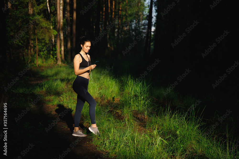 a beautiful athletic Woman in leggings and a t-shirt stands in the forest in the bright sunlight. In the hands of a mobile phone. The girl listens to music with headphones a beautiful athletic Woman in leggings and a t-shirt stands in the forest in the bright sunlight. In the hands of a mobile phone. The girl listens to music with headphones