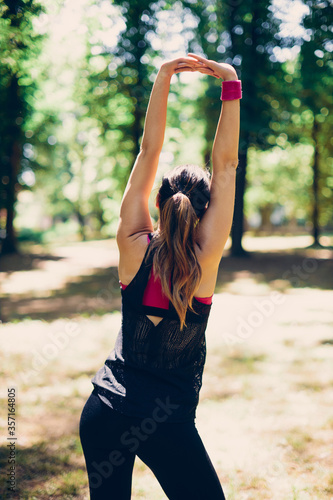 Rear view of attractive caucasian sportswoman standing in nature, stretching backs and arms and preparing for running.