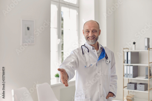 Happy senior doctor reaching out for handshake, welcoming new patient into his medical office.