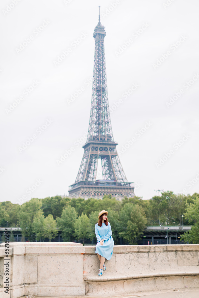 Young woman in blue dress enjoying beautiful landscape view on the Paris and Seine river, while sitting on the bridge on the background of Eiffel tower