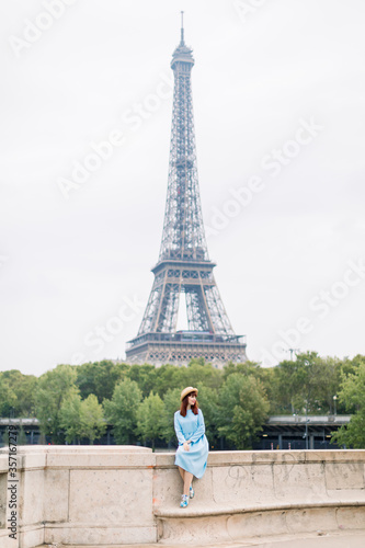 Young woman in blue dress enjoying beautiful landscape view on the Paris and Seine river, while sitting on the bridge on the background of Eiffel tower © sofiko14
