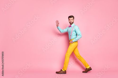 Full length profile photo of handsome guy trend look walk meeting red carpet celebrity waving hand wear shirt suspenders bow tie yellow pants footwear isolated pastel pink color background