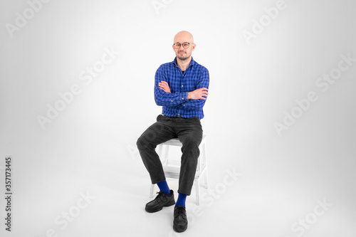 Handsome caucasian man without hair in blue shirt, black trousers and blue socks sits on a long chair and thinks about something isolated on white background photo