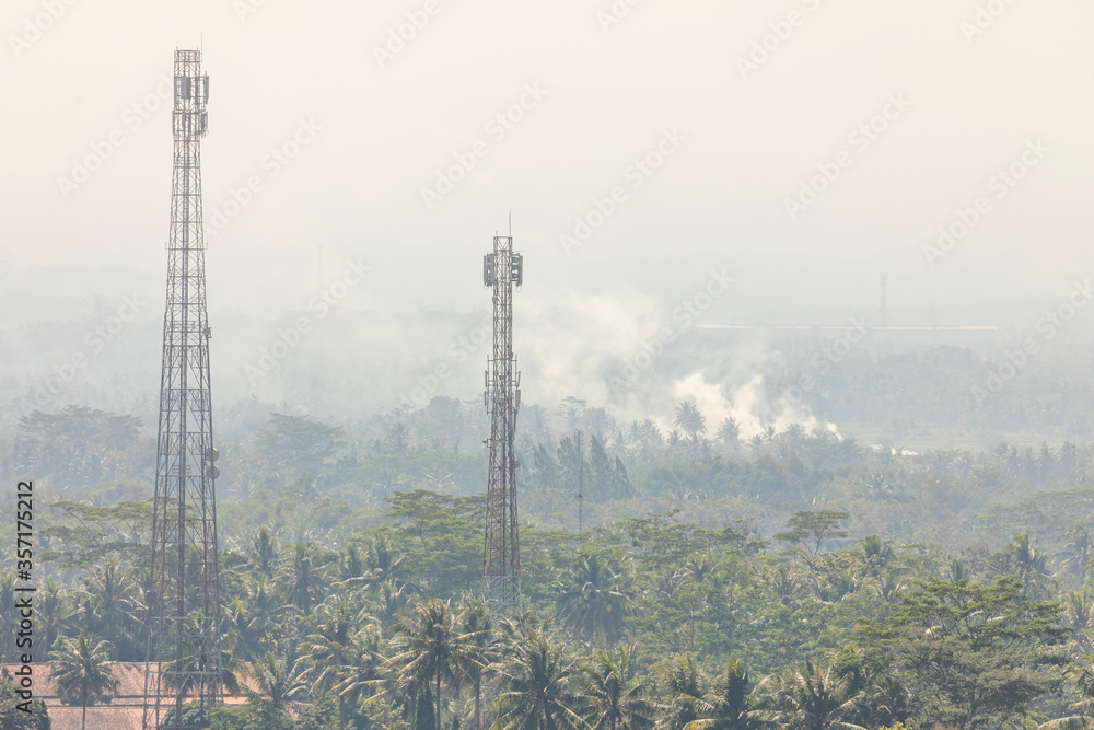 Landscape view of lush palm groves and jungle, with two mobile phone antennas and smoke from some burning in the distance, surrounding the Borobudur temple in Central Java, Indonesia.