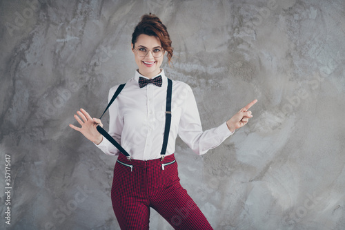 Portrait of her she nice-looking attractive lovely content cheerful cheery wavy-haired girl pointing forefinger ad pulling suspender isolated on gray concrete industrial wall background