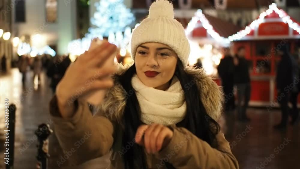 Beautiful smiling girl making selfie with smartphone on the winter street with christmas lights in SLOW MOTION HD VIDEO. Glowing blurred Christmas lights decoration in the background. Beautiful smiling girl making selfie with smartphone on the winter street with christmas lights in SLOW MOTION HD VIDEO. Glowing blurred Christmas lights decoration in the background.