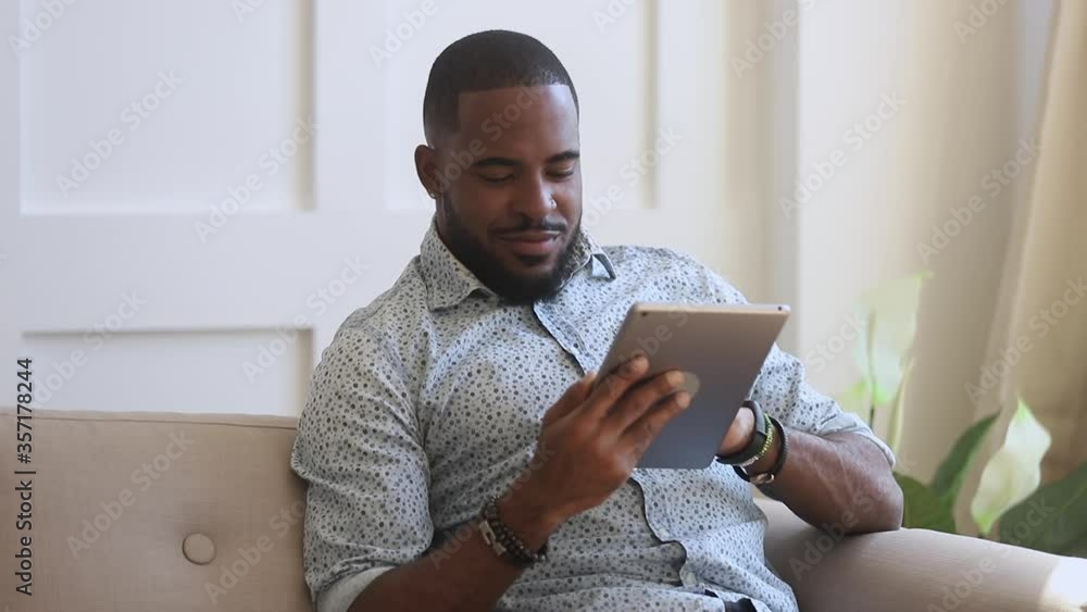 Smiling black man using digital tablet sit on couch at home, young african guy holding computer looking at screen reading e-book app online relaxing on leisure with wireless device work study online