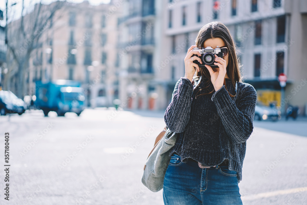 Hipster girl in sunglasses fond of photography using vintage camera strolling on showplaces