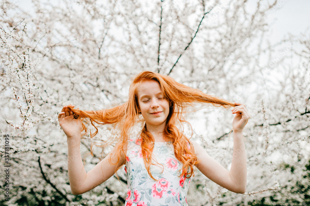 Naklejka premium Picture of an amazing red-haired baby girl in a white dress enjoying a pastime in the big spring garden