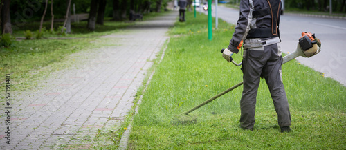 A wide view of the Worker in protective clothing and gloves with a lawn mower in his hands is walking along the lawn nearby along the walking path. A man mows grass with dandelions next to the roadway