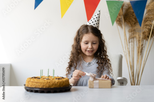 Cute beautiful girl on her birthday. She sits at a table and unpacks a gift. He lies on a table next to a festive cake with candles. The child has loose curly hair and a white cap on his head.