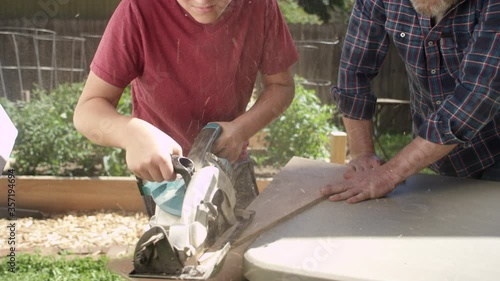 A proud father holds down a piece of wood as his son cuts with a circular saw, they high five