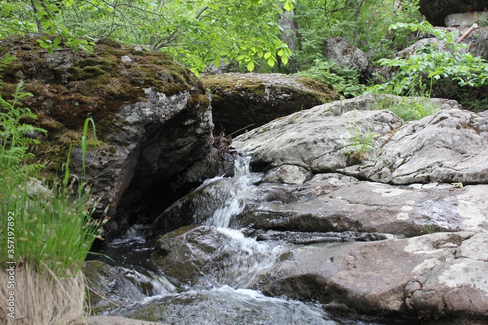 Fototapeta premium Amazing waterfall in the middle of mountain forest. River full of water during spring, beautiful flowing mass. Lovely contrast of water and surrounding forest green. Stones and fresh green leaves. 