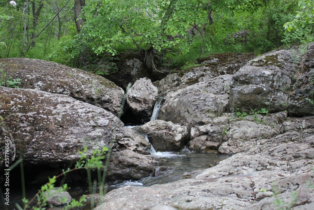 Mountain river - a small waterfall on a river with crystal clear water that flows among gray stones in a green forest on a cloudy summer day. Close up of a rock next to a body of water